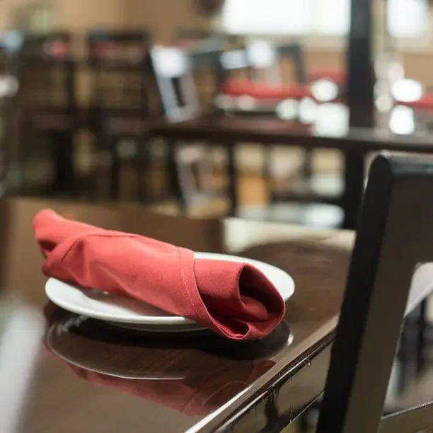 A dining table setting with a white plate topped with a folded red cloth napkin in a restaurant or café.