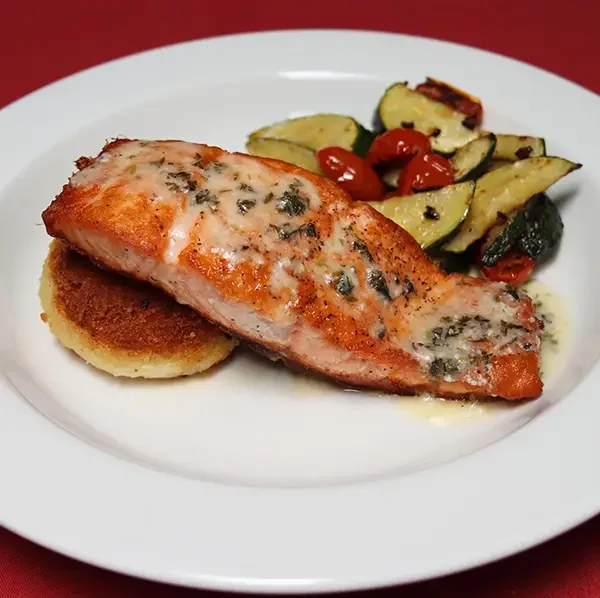 A plate with a cooked salmon fillet topped with a creamy herb sauce, a lemon wedge, a slice of toasted bread, and grilled zucchini and cherry tomatoes.
