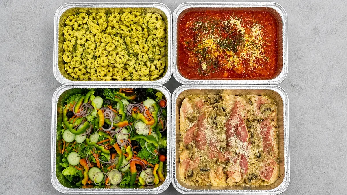 Four aluminum containers with different foods: top left with seasoned pasta, top right with marinara sauce topped with cheese and herbs, bottom left with mixed salad including cherry tomatoes, cucumbers, onions, and greens, bottom right with chicken and ham covered in cheese and olives.