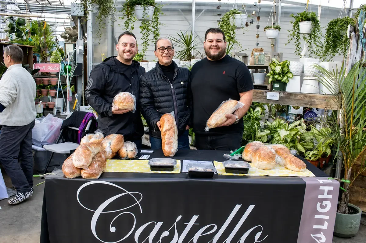 Three men stand behind a table displaying various loaves of bread in a greenhouse filled with plants. They are smiling and holding bread, with more bread on the table and potted plants in the background.