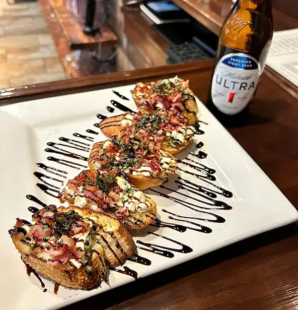 Four slices of bruschetta topped with vegetables, herbs, and bacon, drizzled with balsamic glaze, served on a white plate with a bottle of beer in the background.