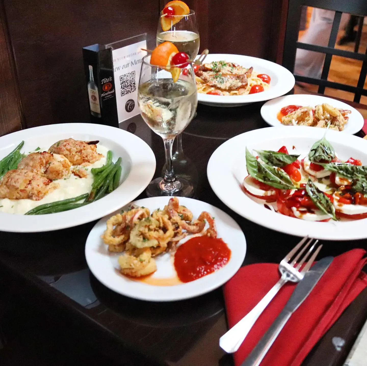 A table set with multiple plates of Italian food, including a Caprese salad with tomatoes, mozzarella, and basil, a plate of fried calamari with red sauce, a dish with green beans and meat, and two glasses of white wine with garnishes. There are utensils on a red napkin and a restaurant menu stand on the table.