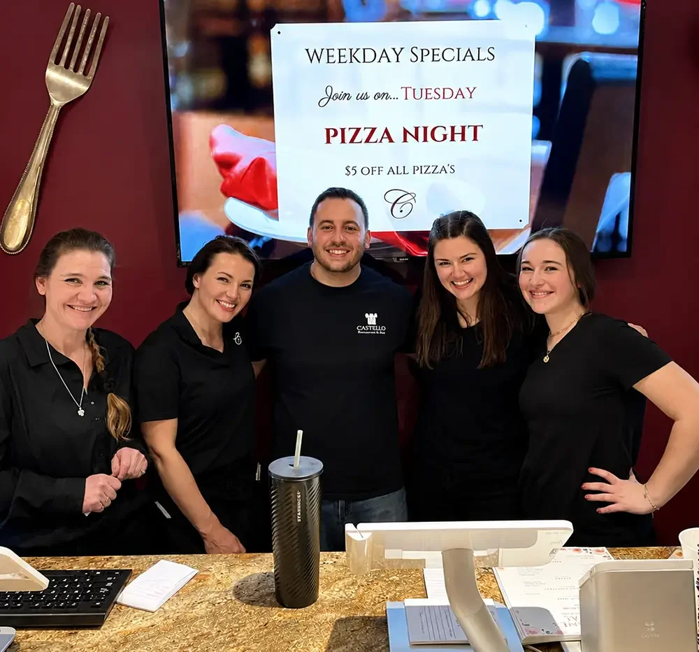 Five staff members standing behind a counter in a restaurant, smiling. Behind them, a screen displays a promotion for Tuesday pizza night with $5 off all pizzas.