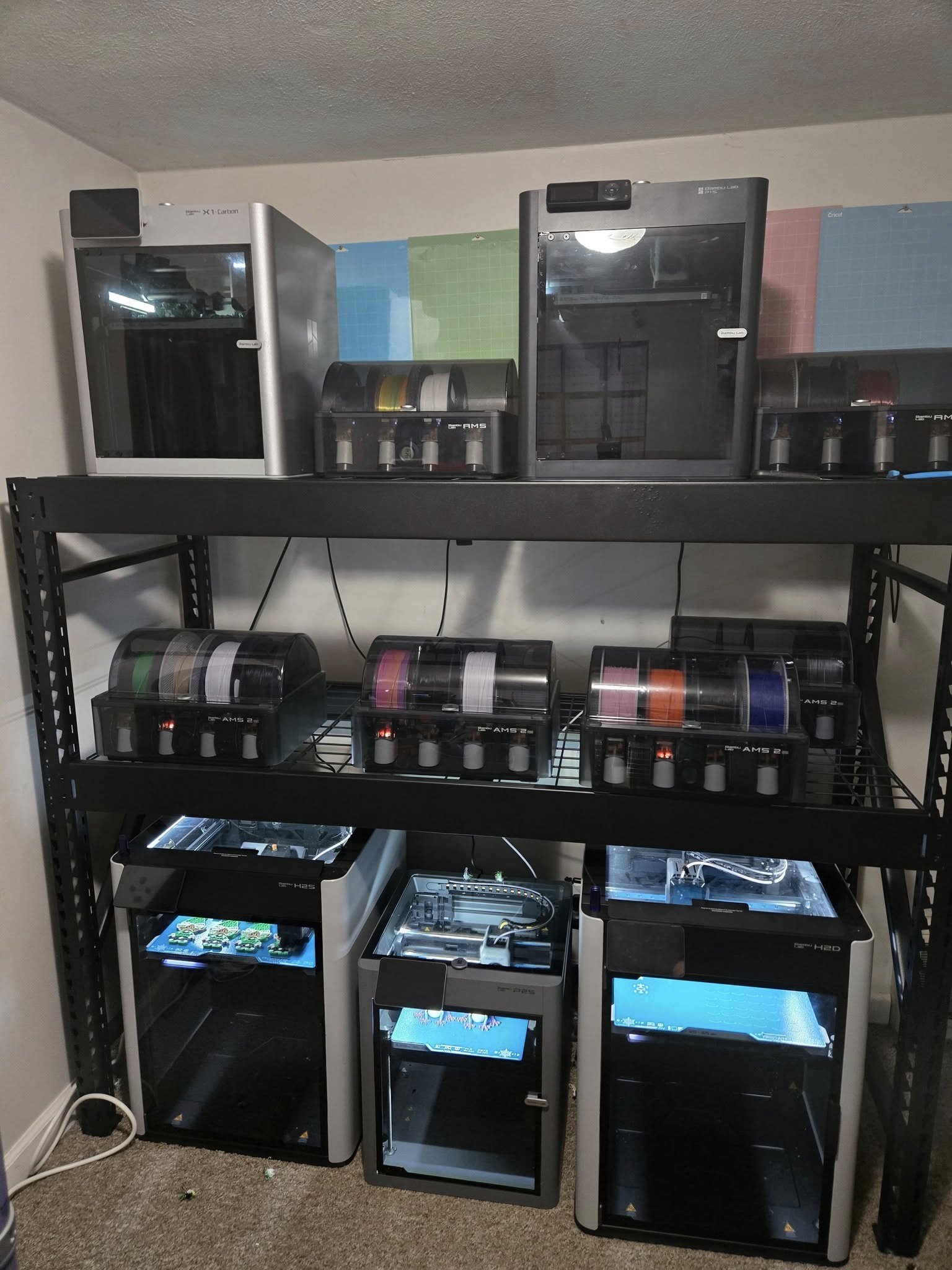 A black metal shelving unit with multiple glass case compartments containing electronic equipment and filament spools against a wall with colored grid paper.