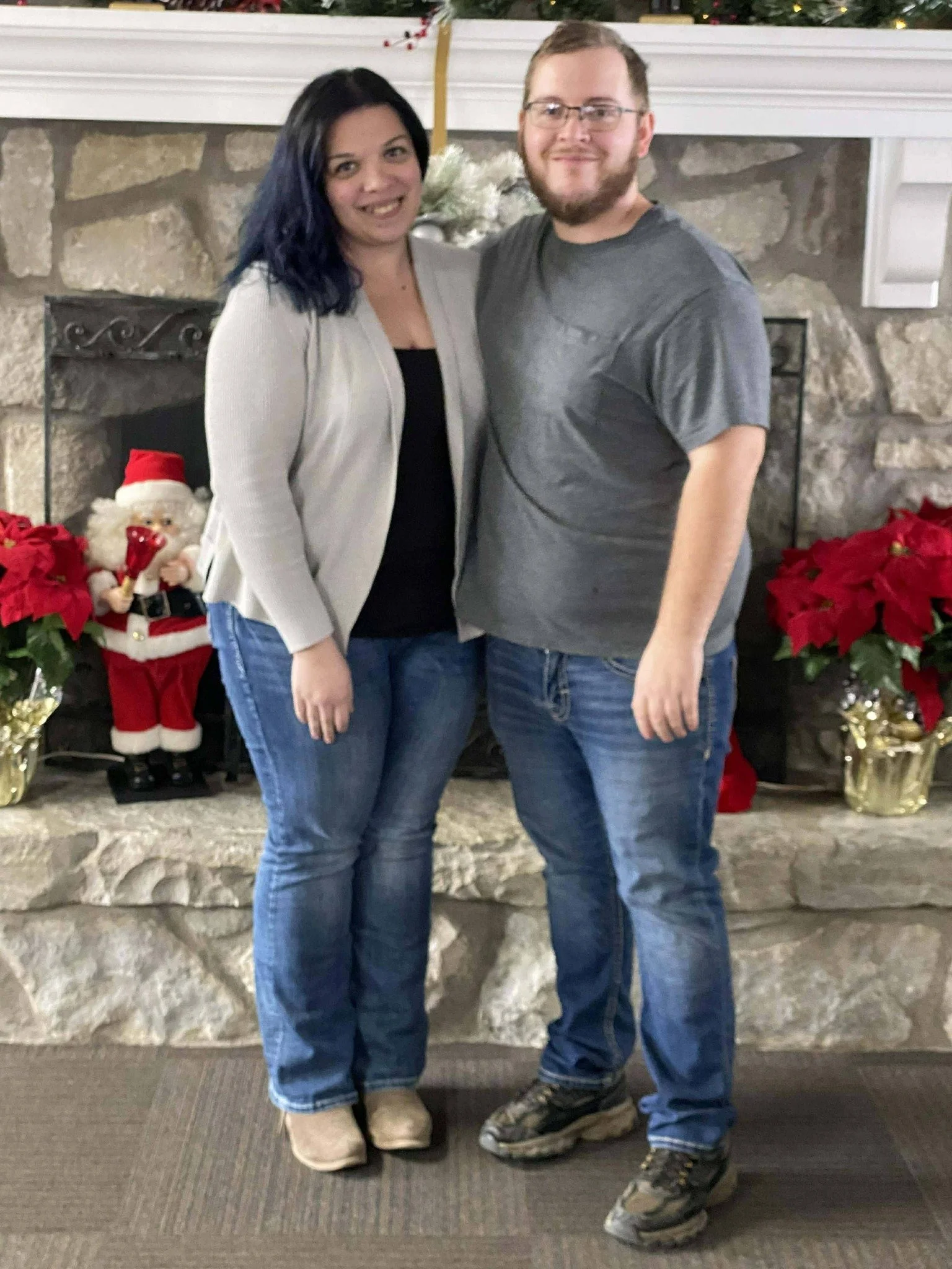 A couple standing in front of a stone fireplace decorated for Christmas with poinsettias and a Santa statue. The woman has dark hair and is wearing a light-colored cardigan and jeans, while the man has a beard, glasses, and is wearing a gray t-shirt and jeans.