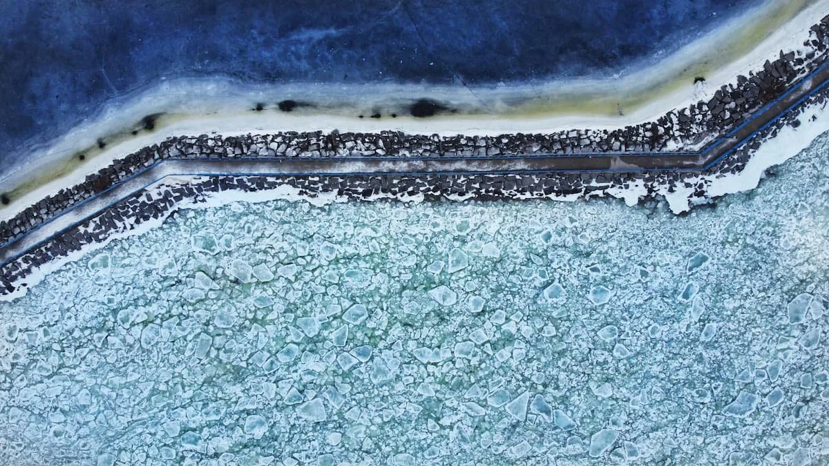 Aerial view of a frozen body of water with large ice chunks and a shoreline with a road and rocky barrier.