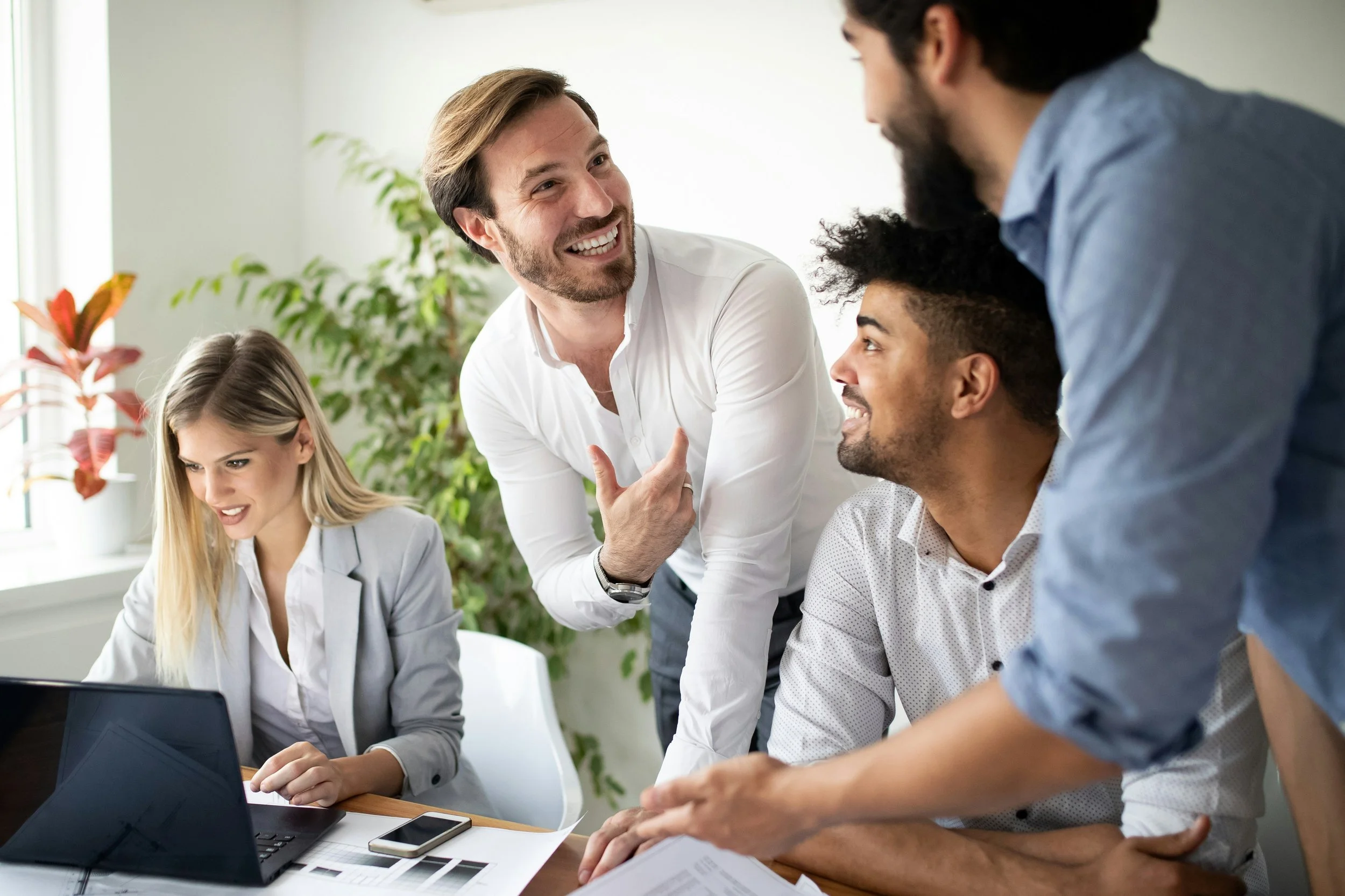 A group of four diverse professionals in an office, engaged in a discussion, with laptops, papers, and a smartphone on the table.