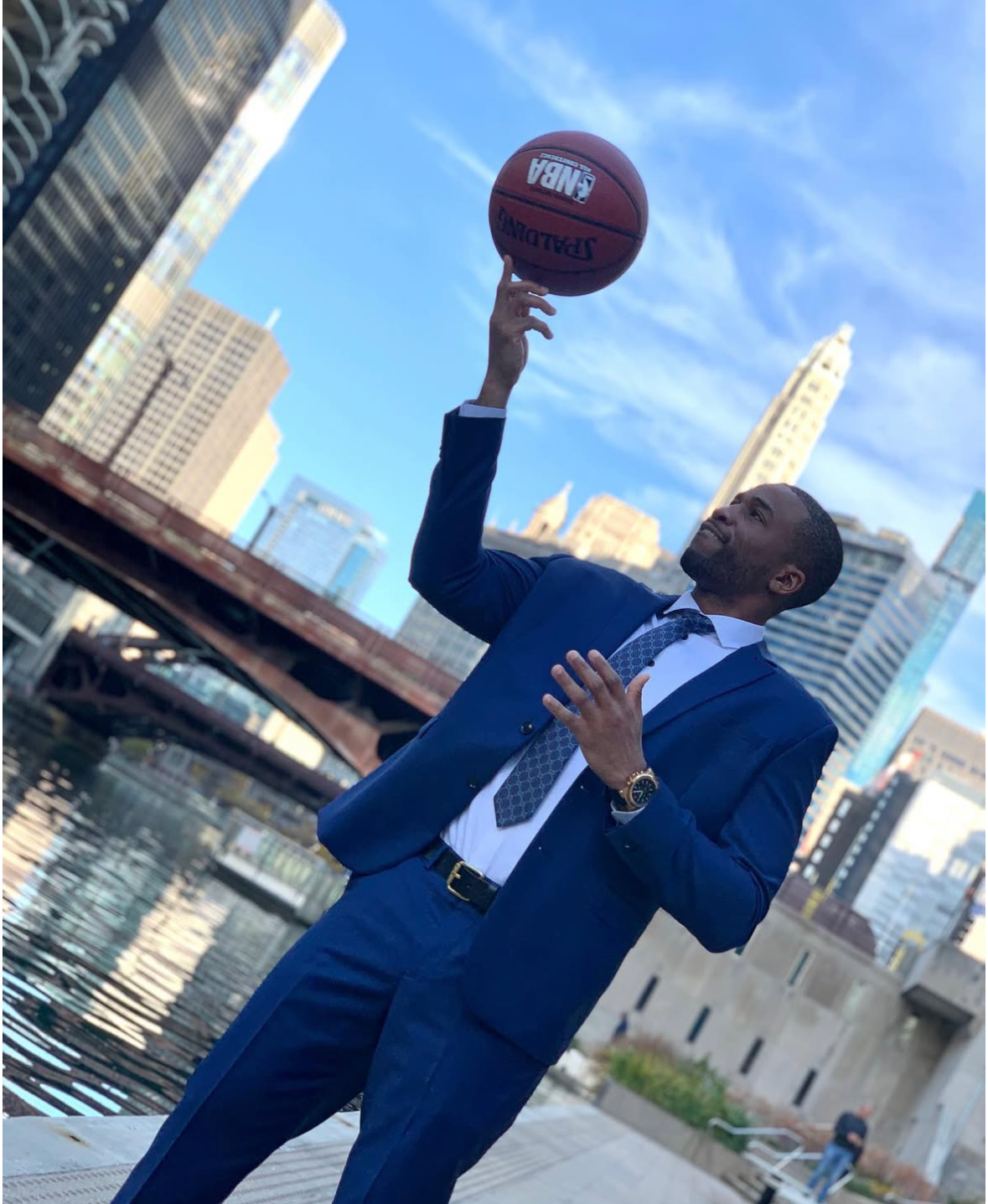 A man in a blue suit holding a basketball in an urban setting with skyscrapers and a bridge.