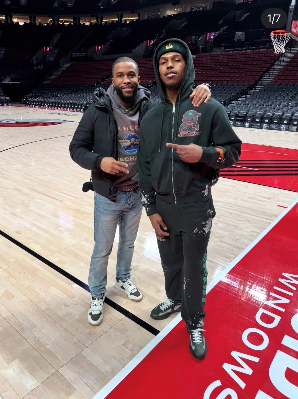 Two young men standing on a basketball court inside a stadium, posing for a photo. The man on the left is smiling, wearing jeans, a grey hoodie, and a black jacket. The man on the right is pointing at his hoodie, wearing black pants, a black hoodie with a graphic print, and a green beanie. The empty stadium seats and basketball hoop are visible in the background.
