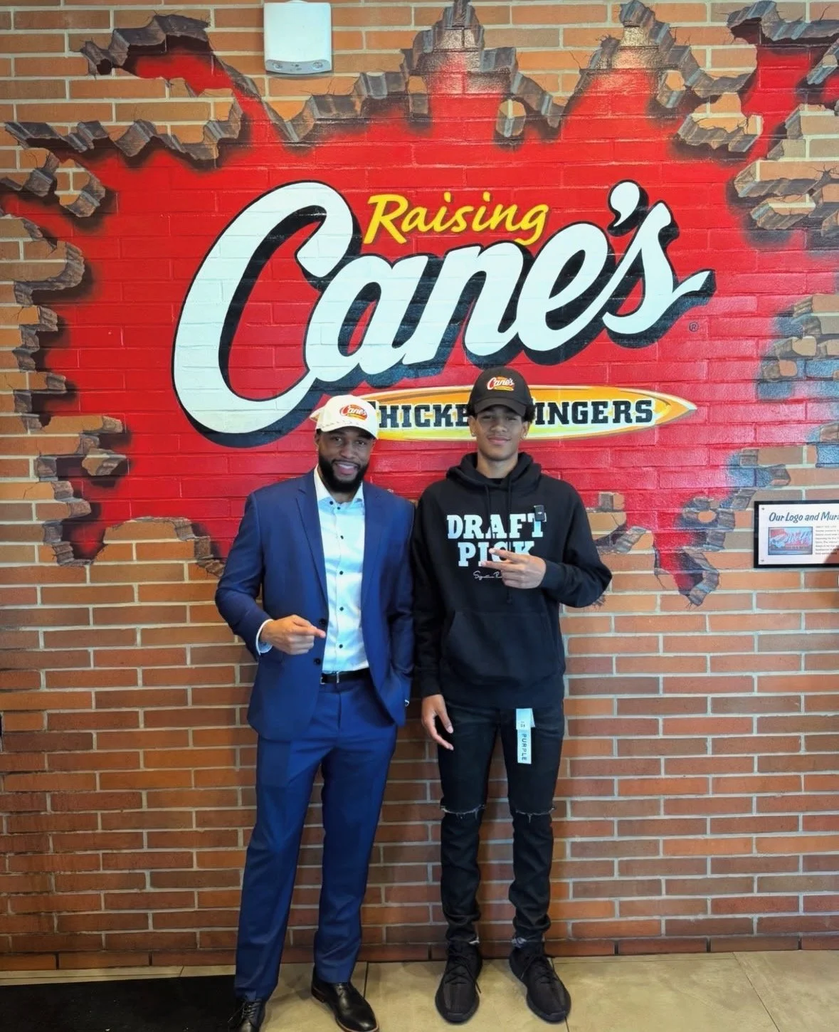 Two men standing in front of a brick wall with a mural that says 'Raising Cane's Chicken Fingers.' One man is dressed in a blue suit and the other in a black hoodie and ripped jeans.