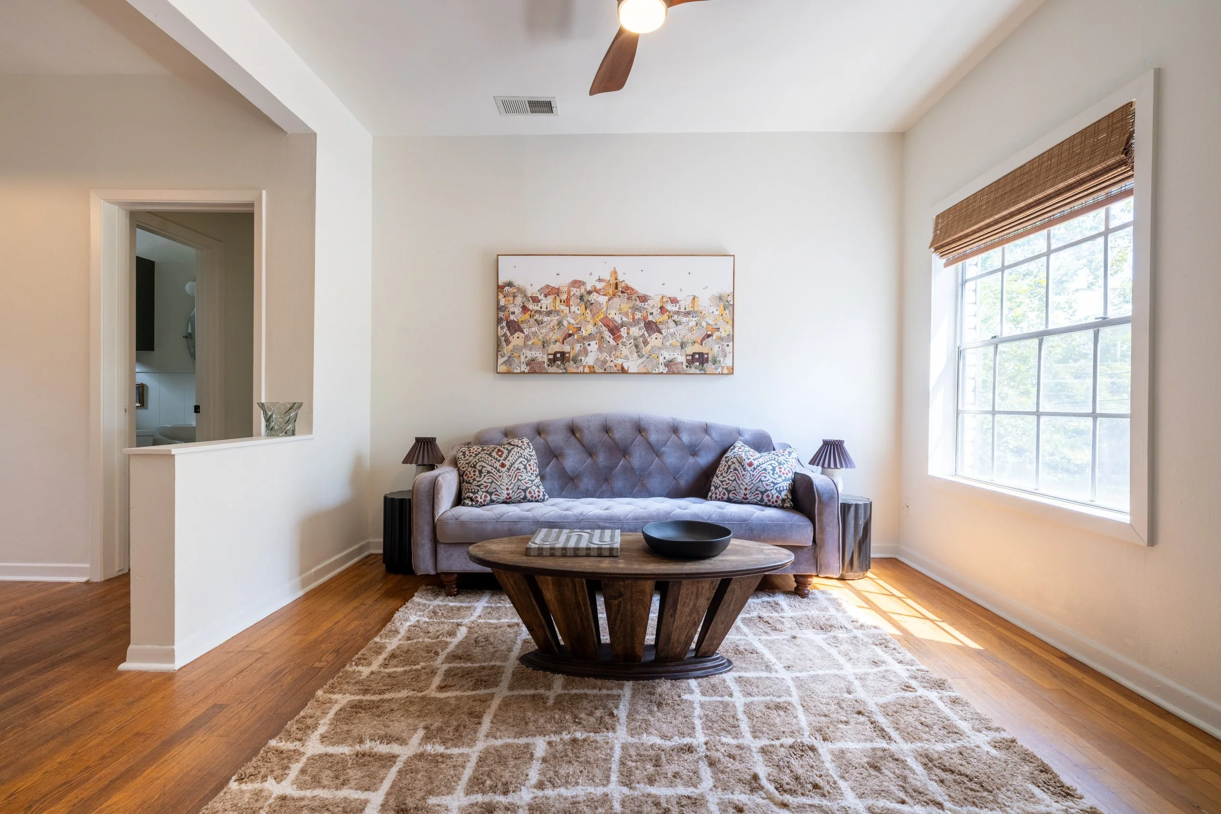 A cozy living room with a gray tufted sofa, patterned pillows, a wooden oval coffee table, two side lamps, a large window with bamboo blinds, a modern framed painting of a townscape, a beige area rug, and a ceiling fan with wooden blades.