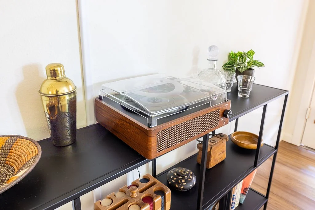 Black metal shelf with a wooden record player and decorative items including a glass decanter, a potted plant, a glass, and wooden bowls.
