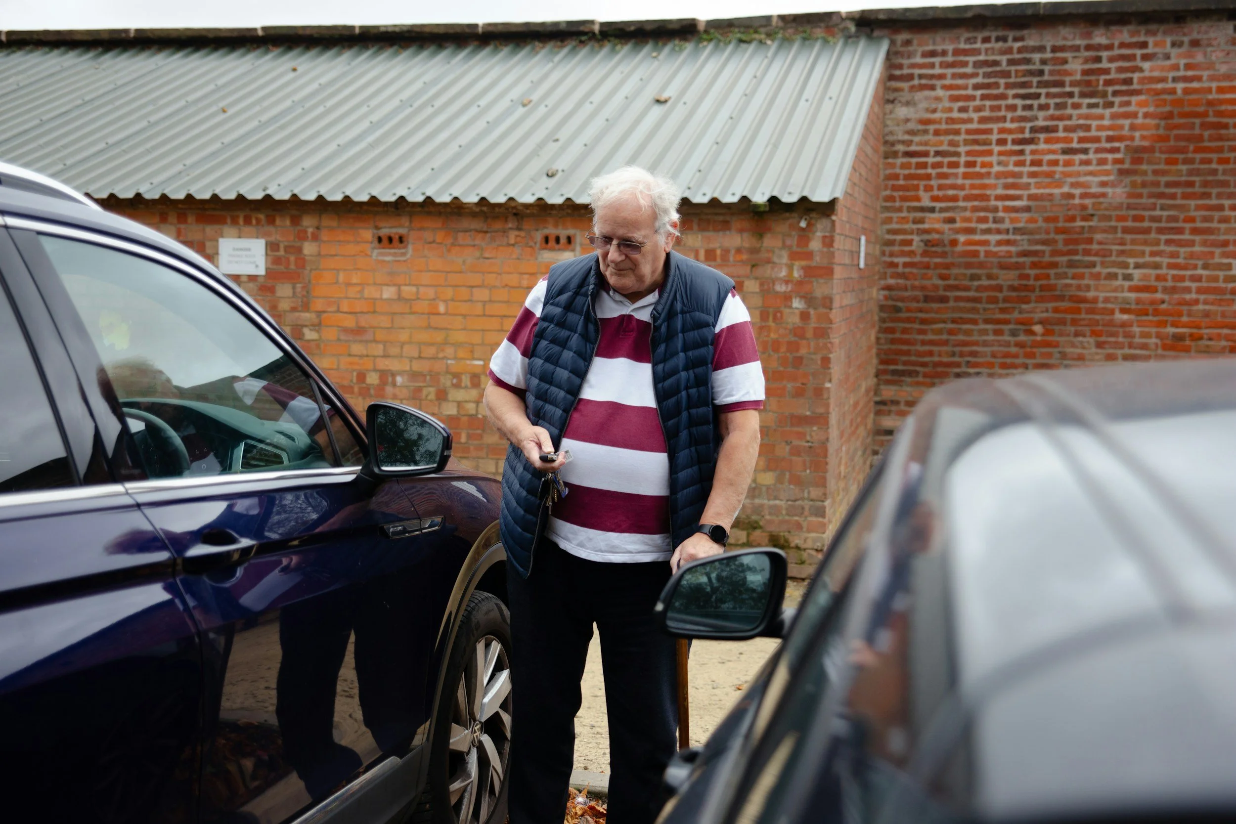 An elderly man with white hair and glasses, wearing a striped maroon and white polo shirt and a navy vest, standing between two parked cars outside near a brick building. He is looking at his phone and holding a set of keys.