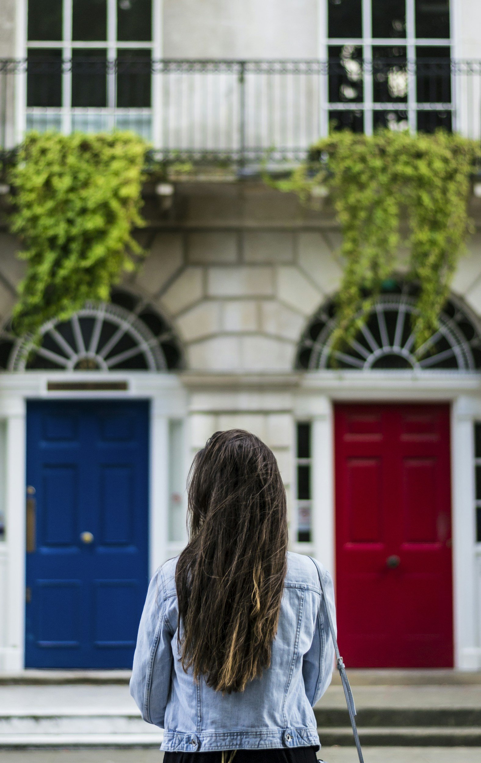 Person with long, dark hair wearing a light blue denim jacket standing in front of a row of houses featuring a blue door and a red door, with green plants and large windows on the houses.