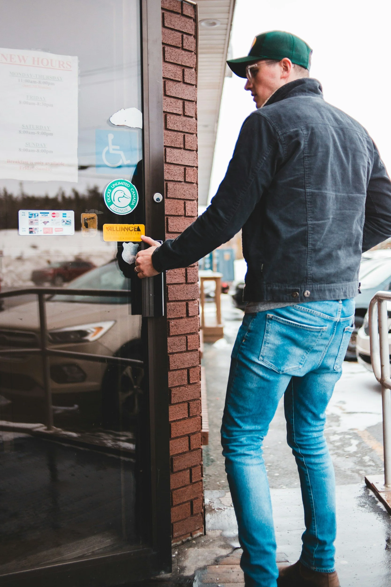 A man wearing a green baseball cap, glasses, a black jacket, and blue jeans is entering a building through a glass door.