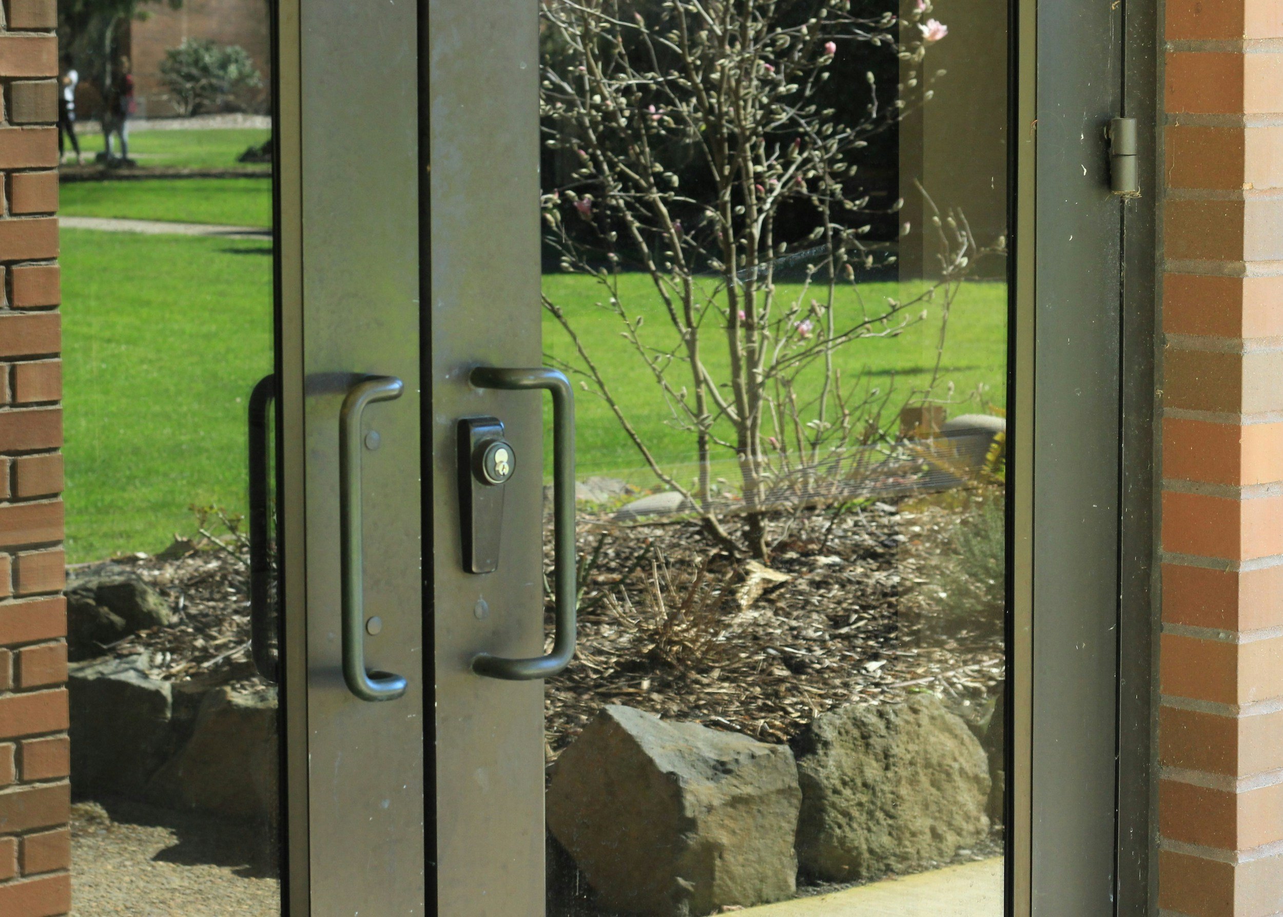 A glass door with metal handles and a keyhole, viewed from the outside, with a small tree or shrub with pink flowers visible behind the door. The surrounding structure is made of brick and metal, and the garden area beyond has grass and other trees.