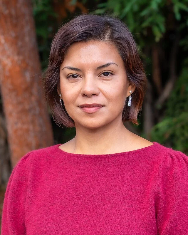 Portrait of a woman with short brown hair, wearing a red top and earrings, standing outdoors with trees and greenery in the background.