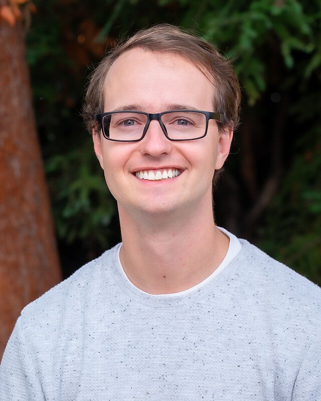 Portrait of a young man wearing glasses, smiling outdoors with trees in the background.