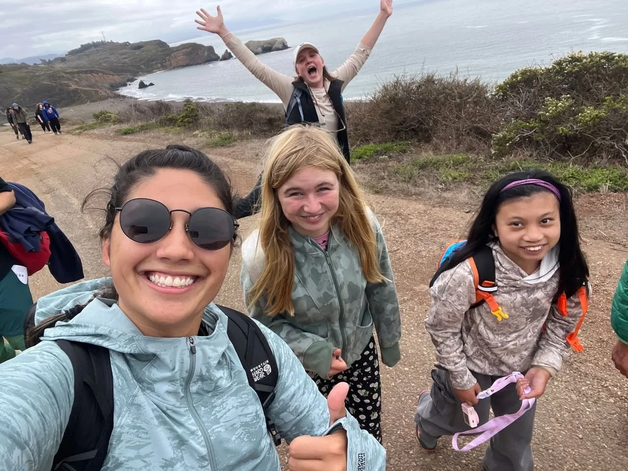 Group of smiling children and a woman hiking on a coastal trail with ocean and cliffs in the background.