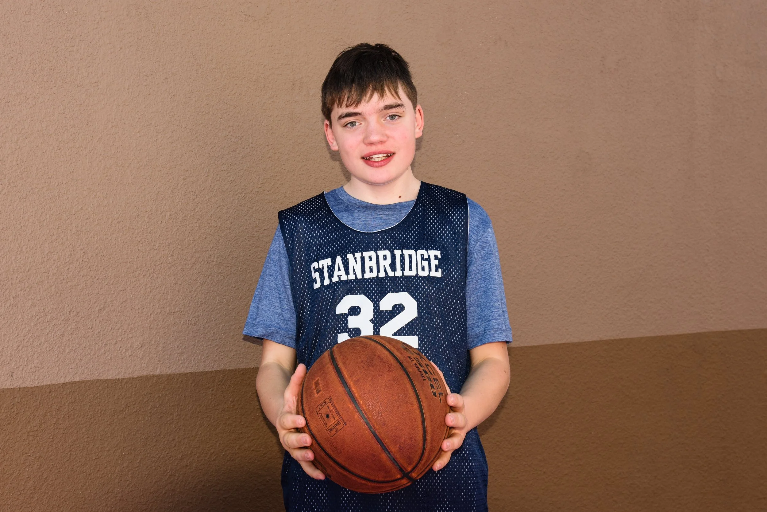 A young boy with short brown hair and light skin is standing against a tan textured wall, wearing a blue sports jersey with the words 'Stanbridge' and the number '32' printed on it. He is holding a basketball with both hands.