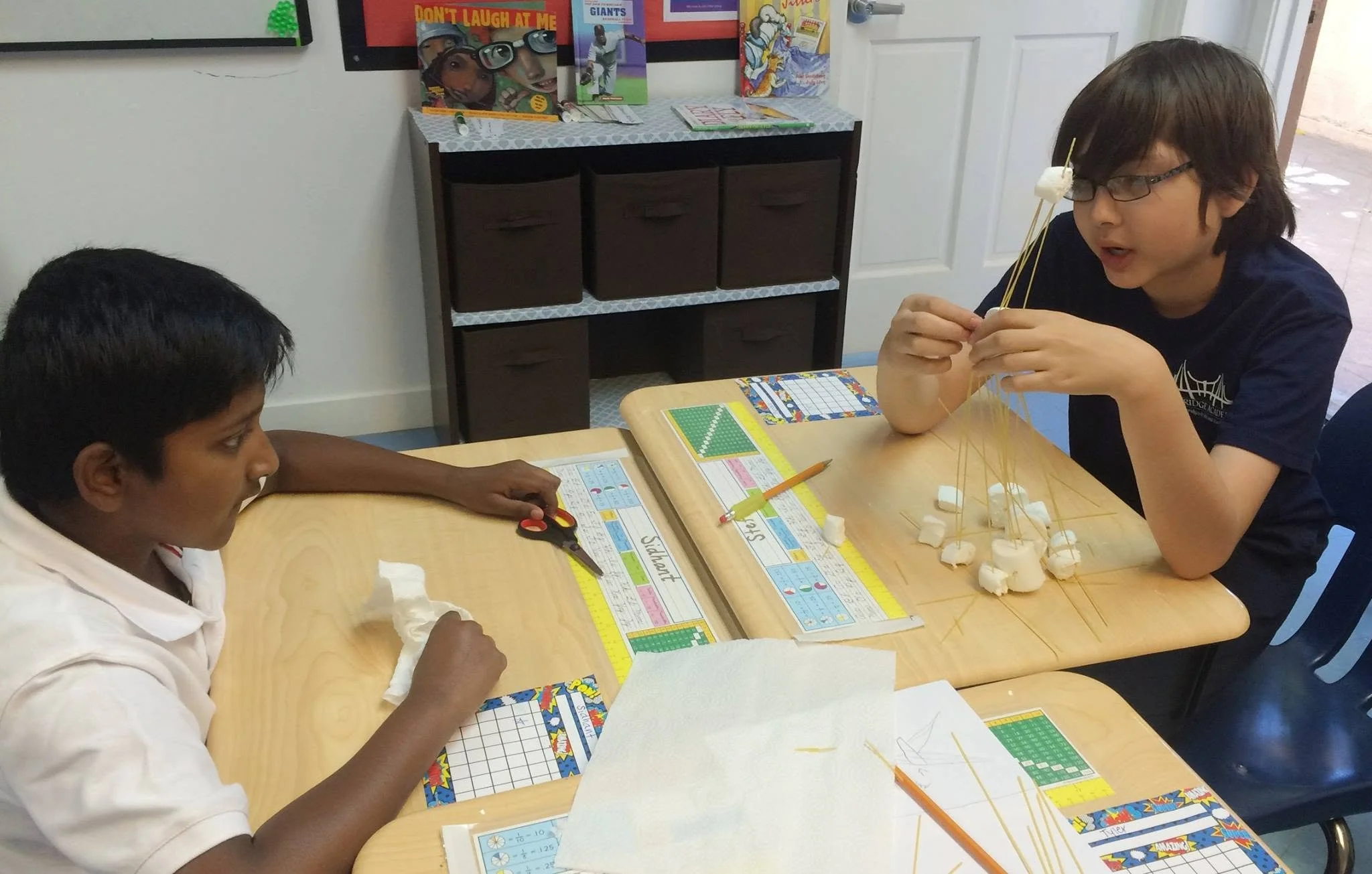 Two children sitting at a table playing a board game involving marshmallows and spaghetti sticks, with educational materials on the table and books on a shelf in the background.