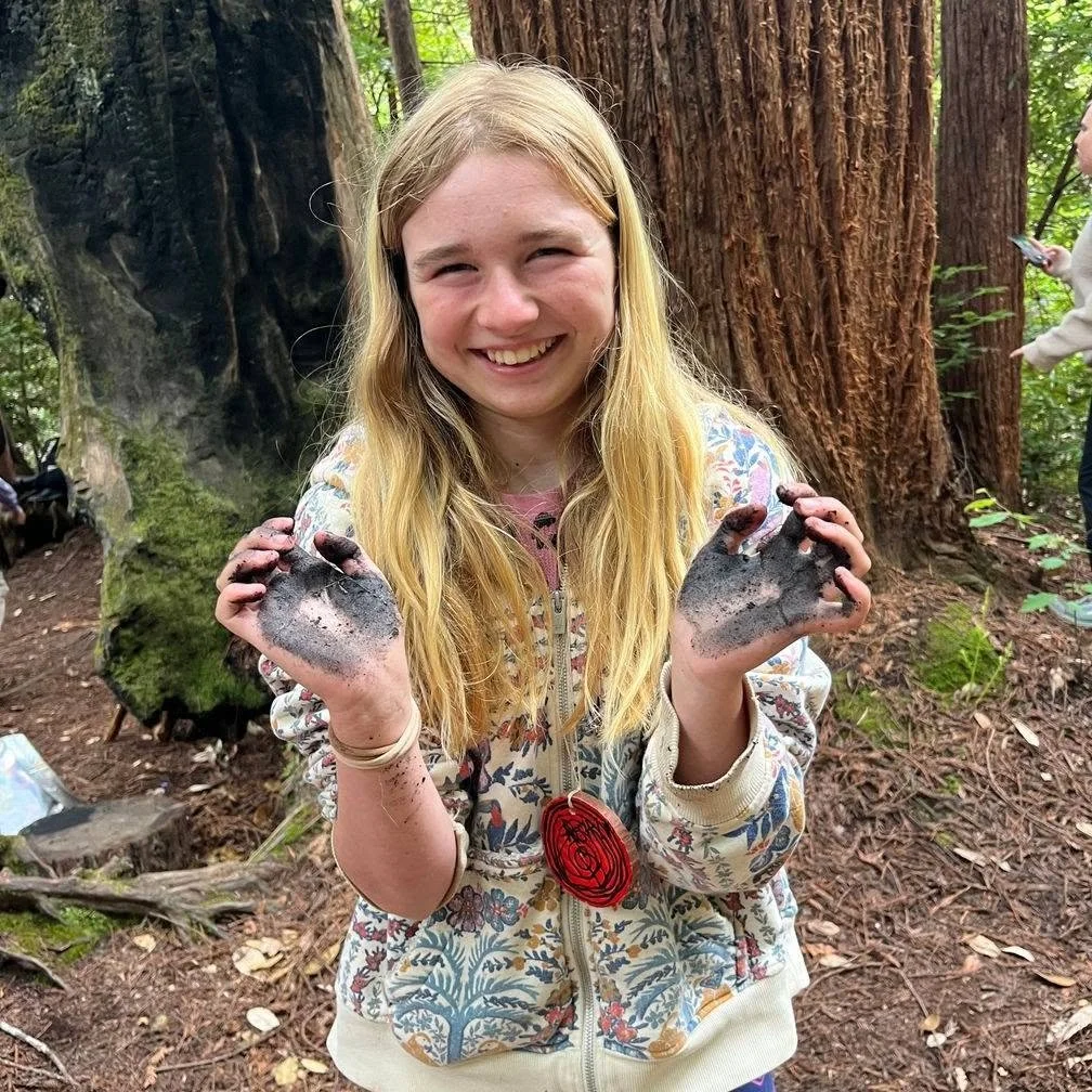 A young girl with long blonde hair smiling and holding muddy hands in a forest setting, with tall trees in the background.