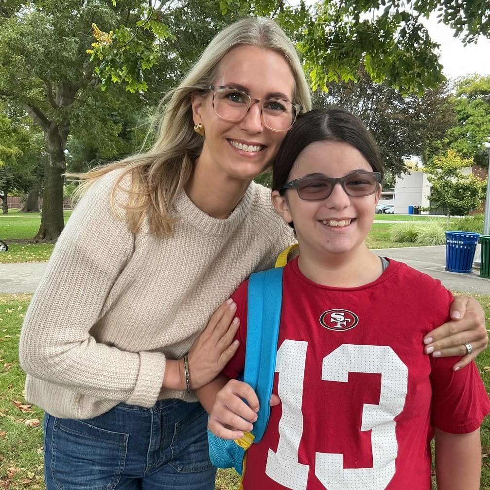 A woman and a girl smiling outside, the woman has blonde hair, glasses, a beige sweater, and blue jeans, and the girl has dark hair, glasses, a red San Francisco 49ers shirt, and a blue backpack.