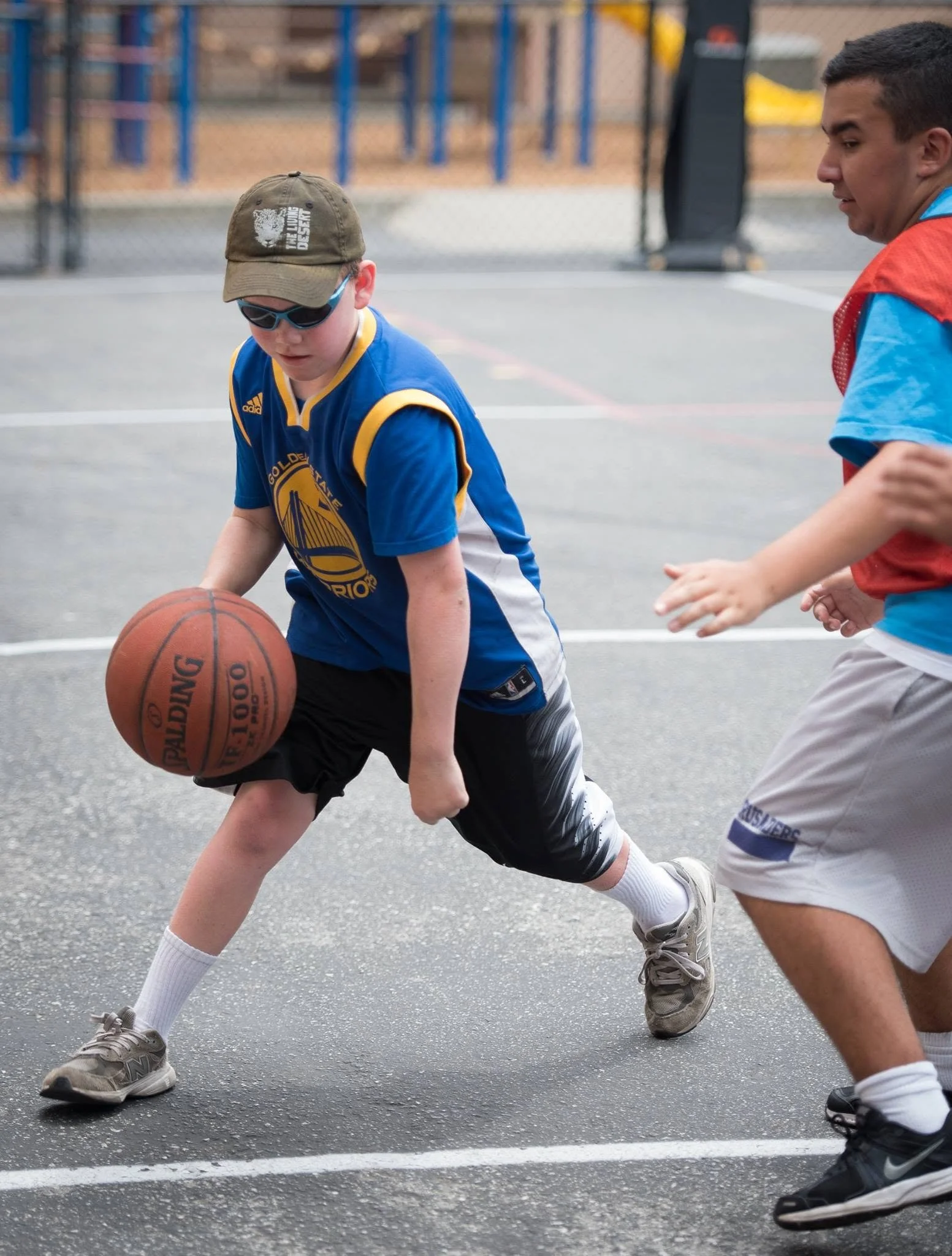 Two boys playing basketball on an outdoor court, wearing athletic clothing and sneakers, with a chain-link fence in the background.