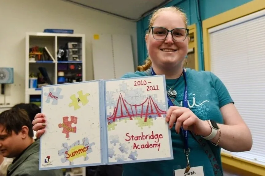 A young woman with glasses and a blue shirt is smiling and holding an open scrapbook or photo album that features a drawing of a bridge, puzzle pieces, and text about Stanbridge Academy from 2010 to 2011.