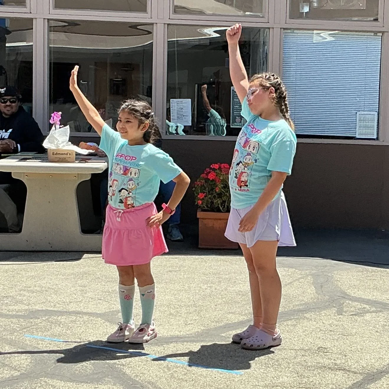 Two young girls standing outside on a sunny day, raising one arm in the air, with a woman standing beside them also raising her arm. They are dressed casually, with colorful t-shirts and skirts, and appear to be participating in an outdoor activity or game. In the background, there is a building with large windows and a seated man at a table.