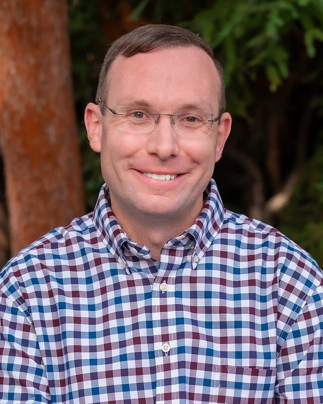A man with glasses smiling outdoors, wearing a checkered shirt, with trees and greenery in the background.