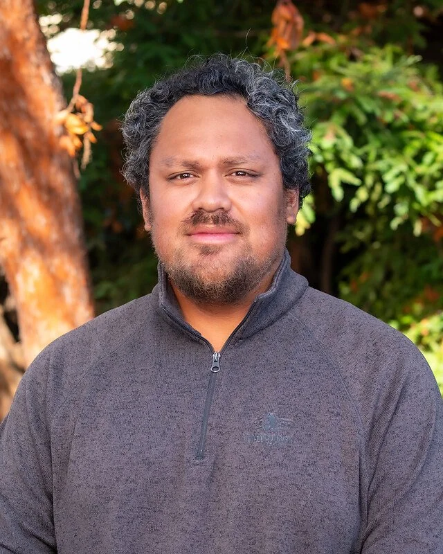 A man with salt-and-pepper curly hair and a beard wearing a dark gray zip-up jacket outdoors with green foliage and a tree in the background.