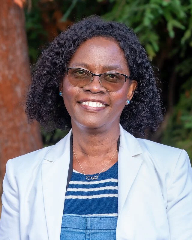 Portrait of a smiling woman with glasses, curly hair, wearing a white blazer and jewelry, outdoors with green foliage in the background.