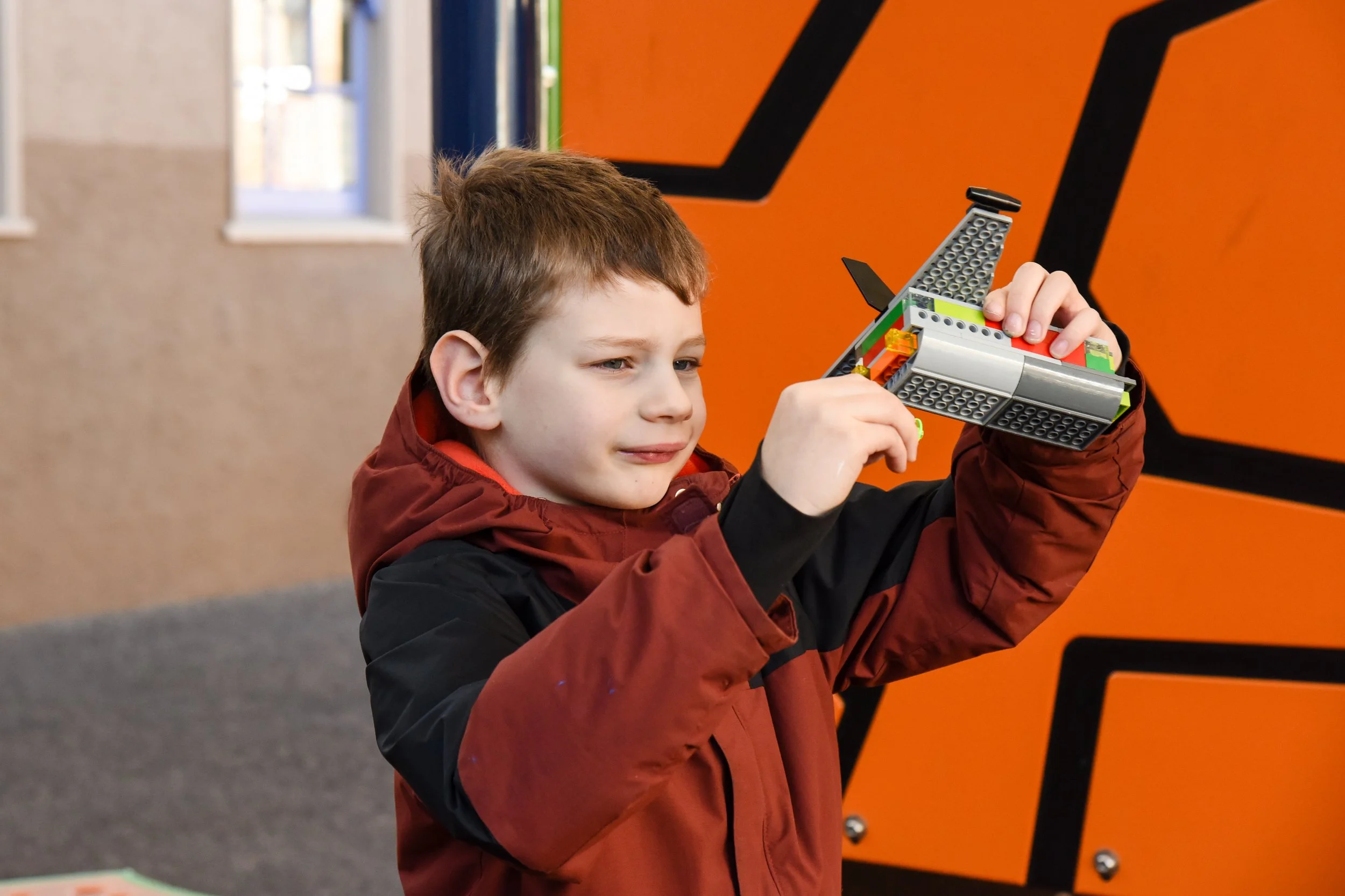 A young boy with short brown hair and a red jacket is holding and examining a LEGO spaceship with a black tilted fin in an outdoor playground setting.