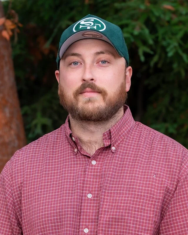 A man with a beard and blue eyes wearing a green baseball cap with a white logo and a pink checkered button-up shirt outdoors with green trees in the background.