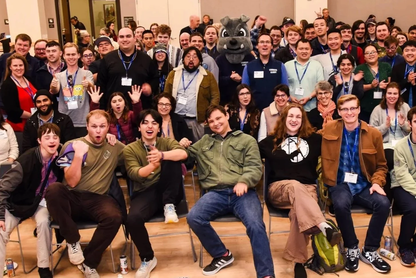 Group of young adults and staff at an alumni reunion, some wearing name tags and a person in a hippo mascot costume in the back. Everyone is smiling, laughing, or waving, with chairs and some backpacks visible.