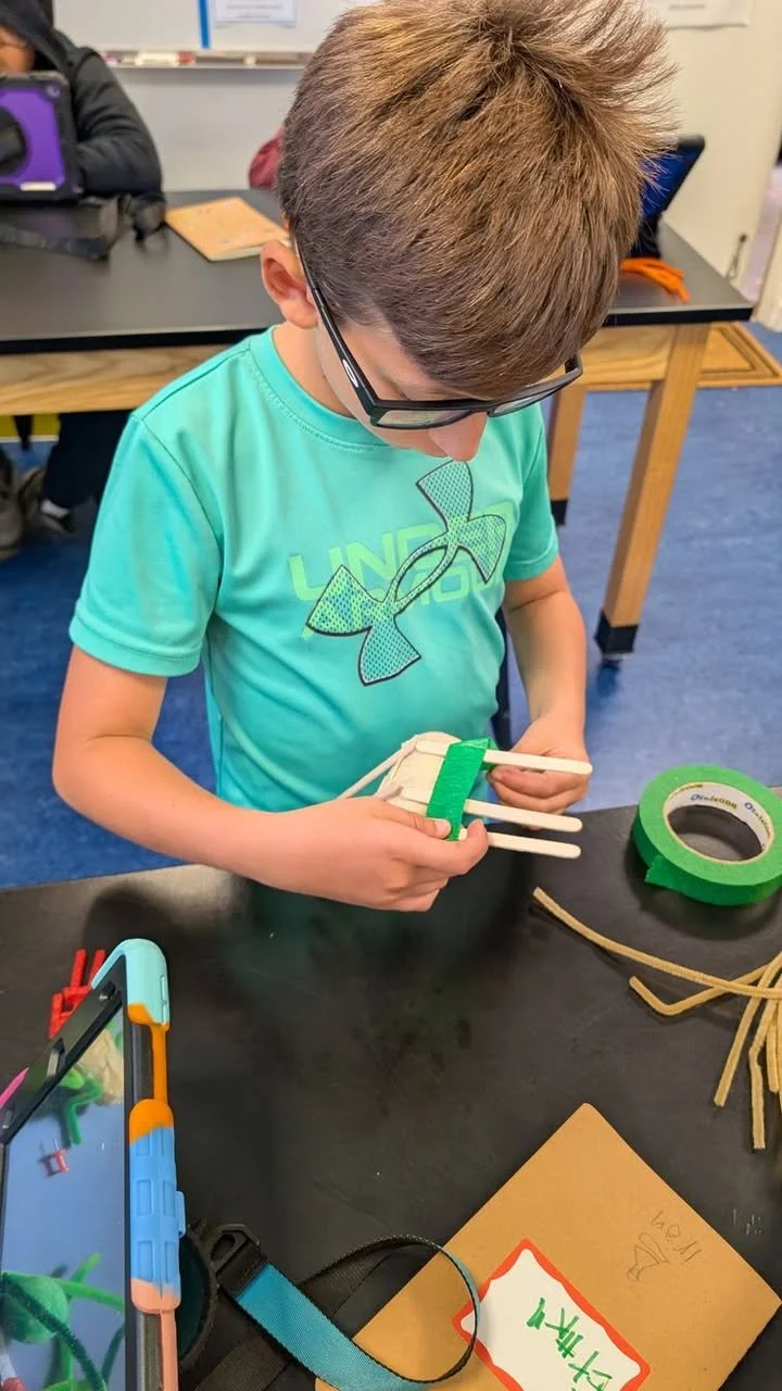 Young boy in a teal T-shirt and glasses working on a craft project with popsicle sticks and green tape at a classroom table. Another person is partially visible in the background.
