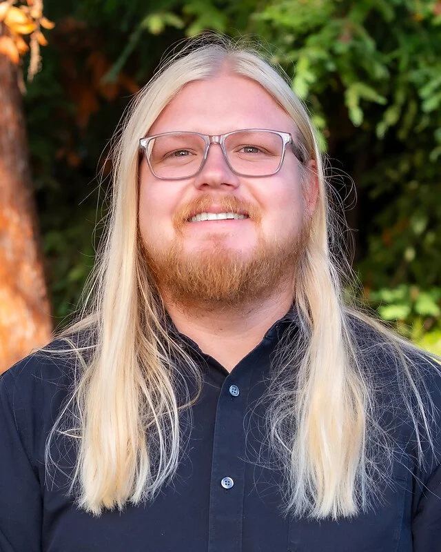 A young man with long blond hair, a beard, and glasses, smiling outdoors with green foliage in the background.