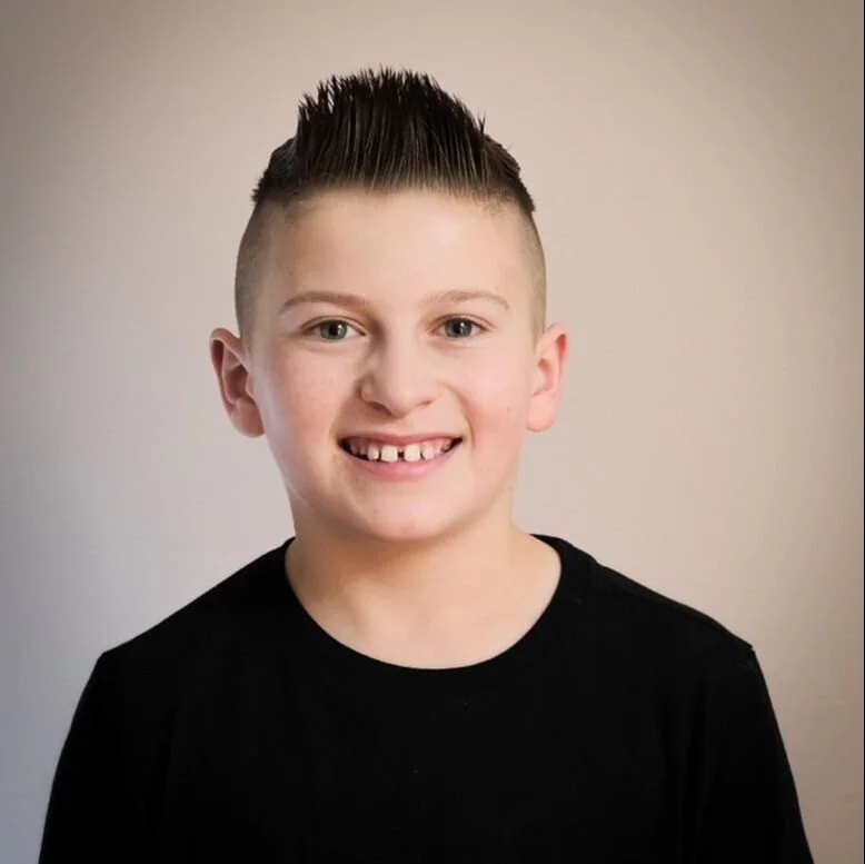 Smiling young boy with a spiked hairstyle, wearing a black shirt against a plain background.