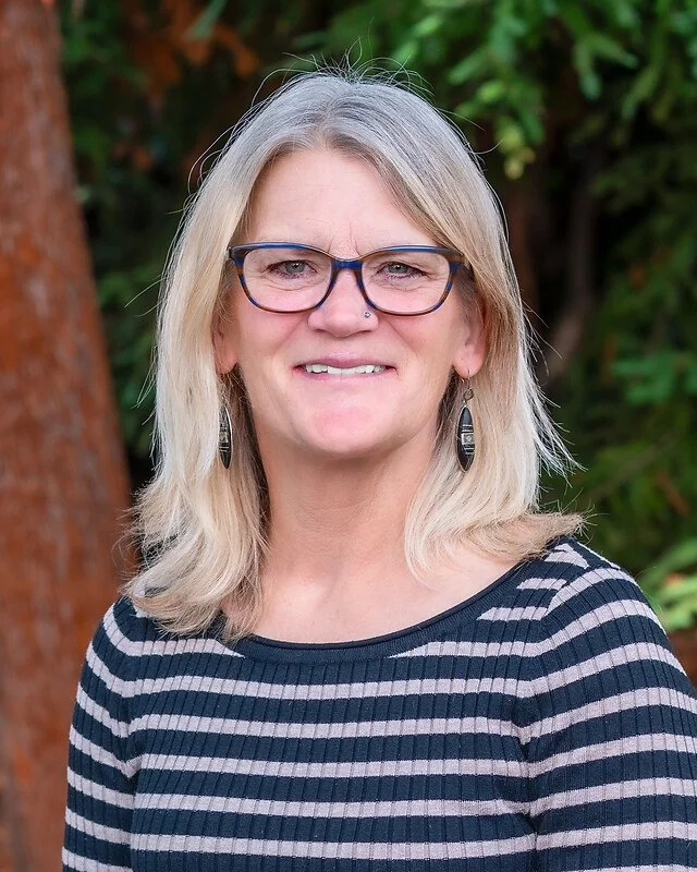 A woman with blonde hair, glasses, and earrings standing outdoors with greenery in the background.