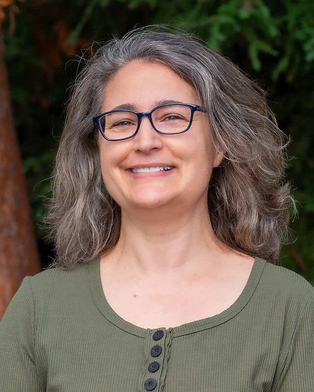 A woman with gray, wavy hair and glasses is smiling outdoors with a background of trees and greenery.