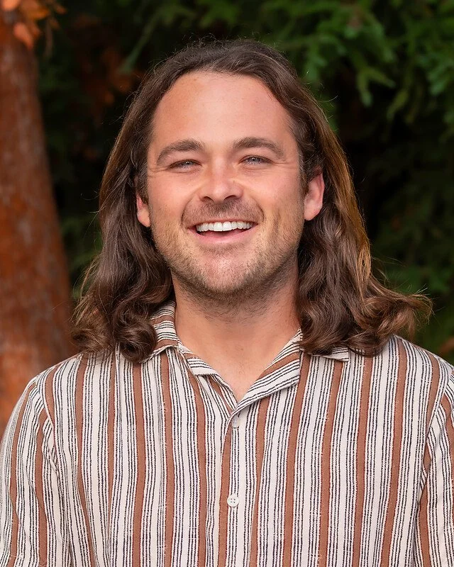 A smiling man with long, wavy brown hair, wearing a striped shirt, standing outdoors with trees and greenery in the background.