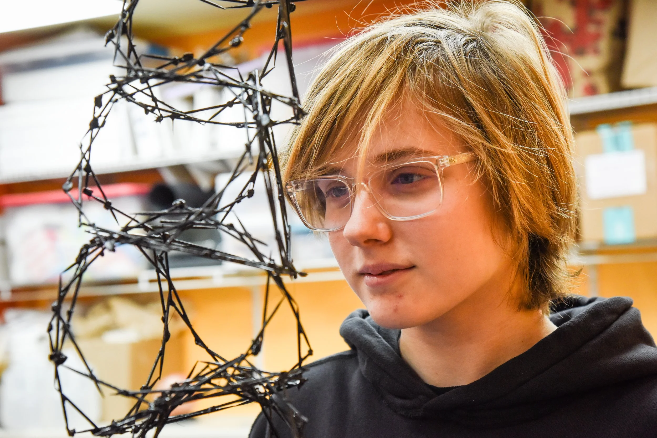A young person with light-colored hair and glasses examining an abstract black wire sculpture in an art room setting.