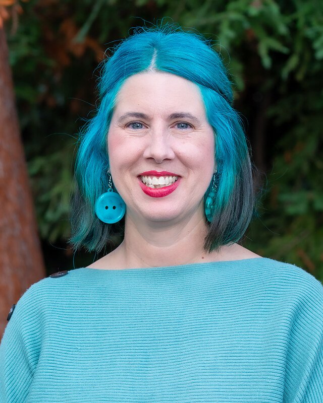 Woman with blue and black hair, bright blue top, large blue button earrings, smiling outdoors with green foliage background.