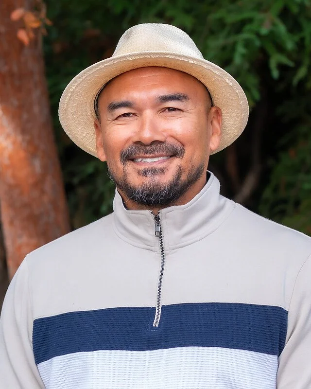 A man smiling outdoors, wearing a straw hat and a zip-up jacket with blue and white stripes, with green foliage in the background.