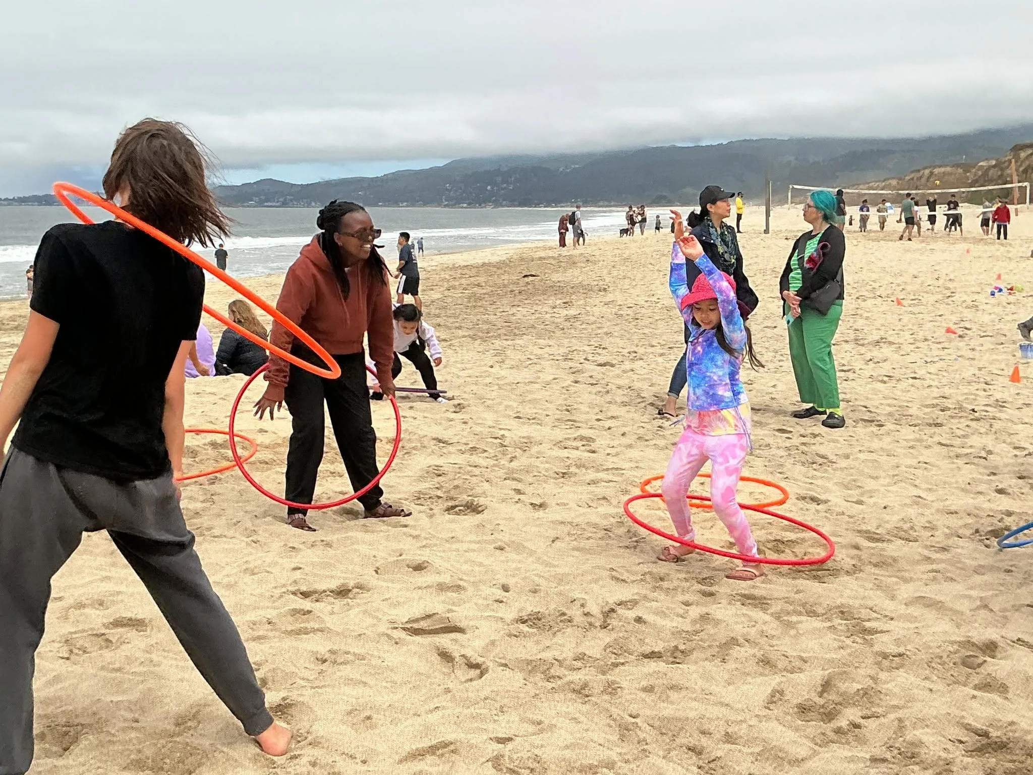 People playing hula hoop games on a sandy beach with waves and mountains in the background.