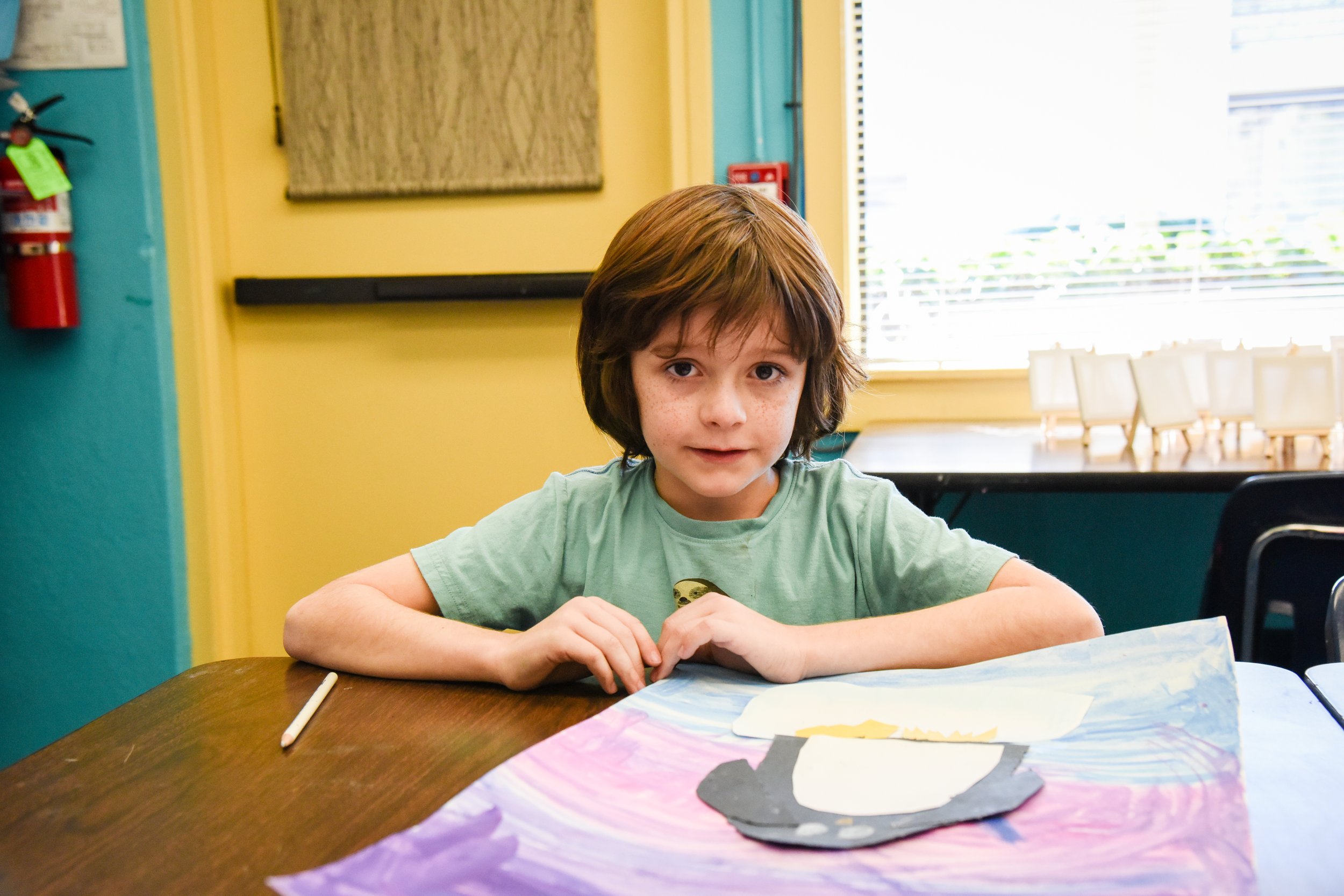 A young boy with brown hair, wearing a light green T-shirt, sitting at a table in a brightly colored room with yellow walls and yellow window trim, working on a colorful art project with paper cutouts.