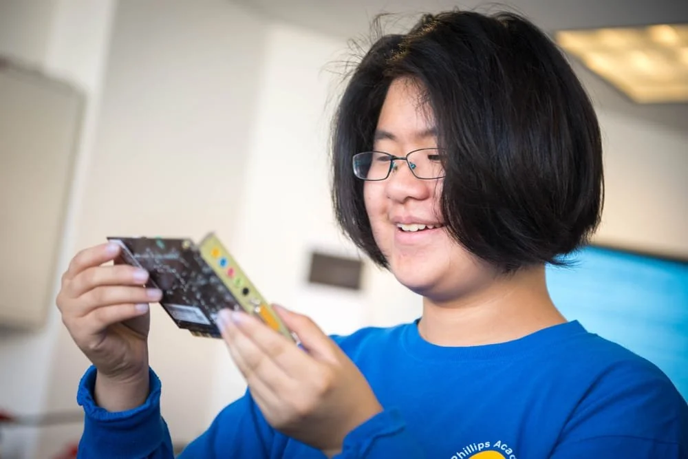 A young person with glasses and straight black hair smiling and looking at a circuit board or electronic device in their hands.