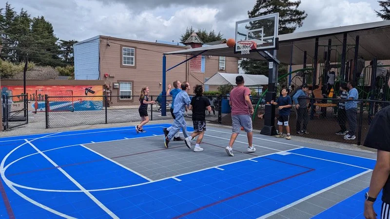 Kids playing basketball on an outdoor court with a colorful mural, fenced area, and spectators nearby.