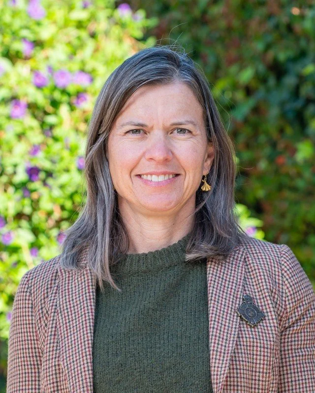 A woman with shoulder-length gray hair smiling outdoors, wearing a checkered blazer over a dark green sweater, with a pin on her blazer and earrings with a small charm.